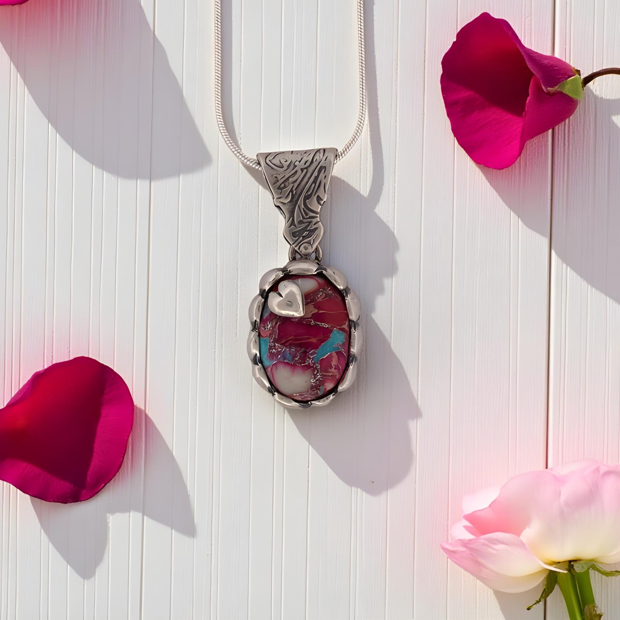 Silver pendant with a pink stone and blue accents on a white background with pink flowers.