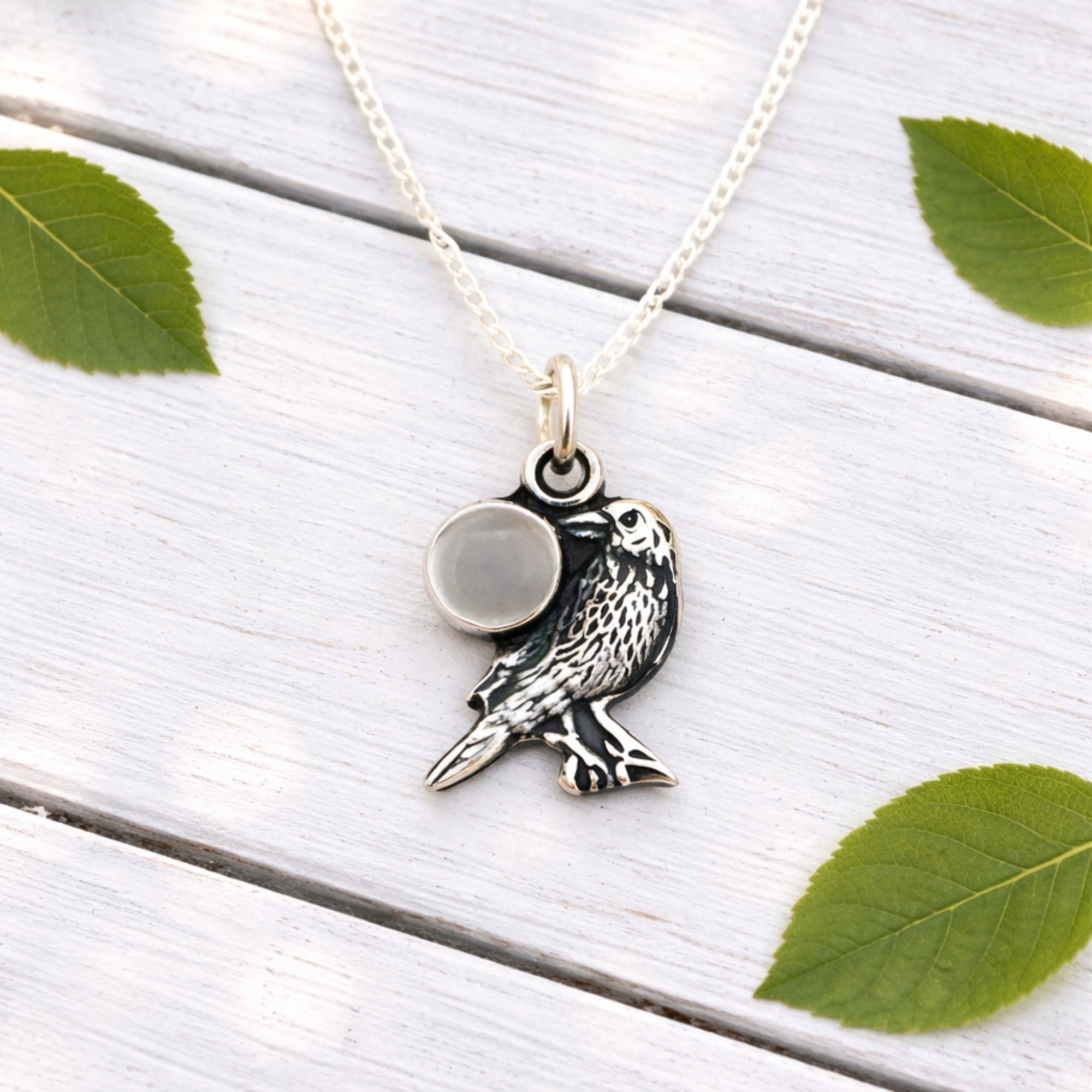 Silver owl pendant necklace with a moonstone on a white wooden surface with green leaves.
